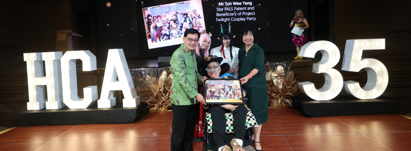 Group with cosplayers and HCA signage & lit "35" on stage. Man in wheelchair holds framed photo.